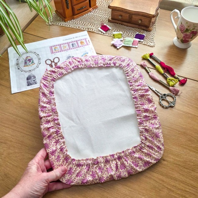 Vue d'ensemble en prise en main d'un grime guard housse protège-toile modèle Samba en tissu coton à motifs floraux rose et violet, ajusté sur un cadre rectangulaire de type Nurge, avec ciseaux vintage, échevettes de fils et grille de broderie sur fond d'a