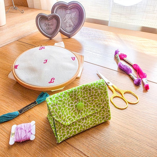 Vue de dessus d'un ensemble coordonné de broderie modèle Verveine comprenant un grime guard installé sur cadre Q-Snap carré et sa poubelle à fils de table assortie, en tissu vert anis aux motifs de feuillage blanc, avec ciseaux dorés, échevettes et boîte 