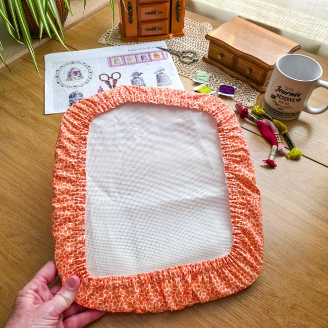 grime-guard-nurge-mandarine-point-de-croix-angelique-la-fee-en-fil Vue en prise en main d'un grime guard housse protège-toile modèle Mandarine en tissu coton orange à motifs gouttelettes blanches, ajusté sur un cadre rectangulaire de type Nurge, avec grille de broderie, échevettes de fils et mug sur une table en bois, cr