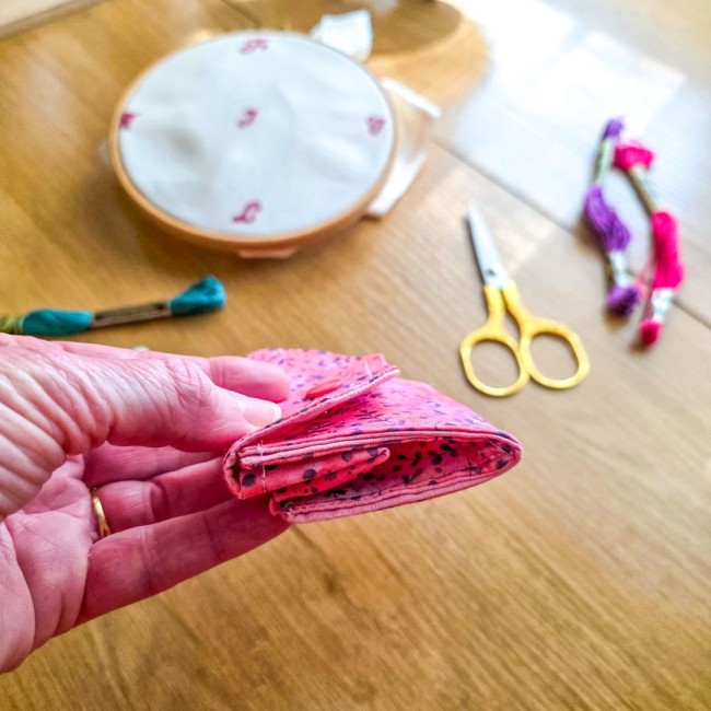 Gros plan de la mini poubelle à fils pliable fermée modèle Sandy Rose tenue en main, montrant sa finesse et sa taille compacte en tissu rose à motifs de feuillage violet, avec tambour à broder, ciseaux dorés et échevettes en arrière-plan, création artisan