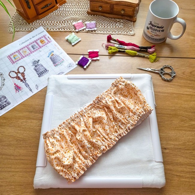 grime-guard-broderie-sous-bois-dore-tissu-beige-replie-qsnap-angelique-la-fee-en-fil Grime guard modèle Sous-Bois Doré en tissu beige plissé à motifs de feuillage ocre, posé au centre d'un métier Q-Snap sur une table de broderie par Angélique La Fée en Fil.