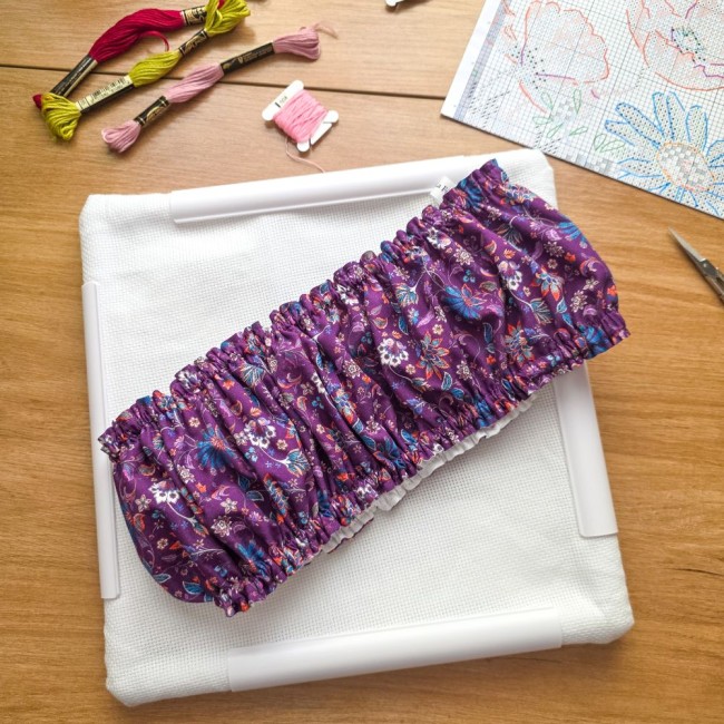 Vue de dessus d'un grime guard housse protège-toile modèle Cachemire en tissu violet aux motifs floraux et cachemire multicolores, posé en travers d'un cadre Q-Snap carré blanc avec échevettes de fils et grille de broderie, création artisanale Angélique L