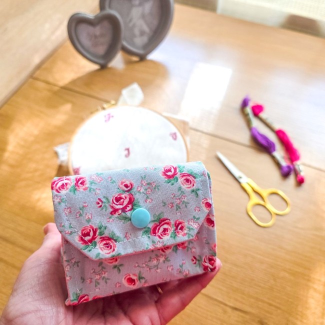 Vue de face d'une mini poubelle à fils pliable fermée modèle Bianca tenue en main, montrant le rabat et le bouton-pression bleu clair sur tissu gris clair aux motifs bucoliques de roses rouges, avec ciseaux dorés et tambour à broder en arrière-plan, créat