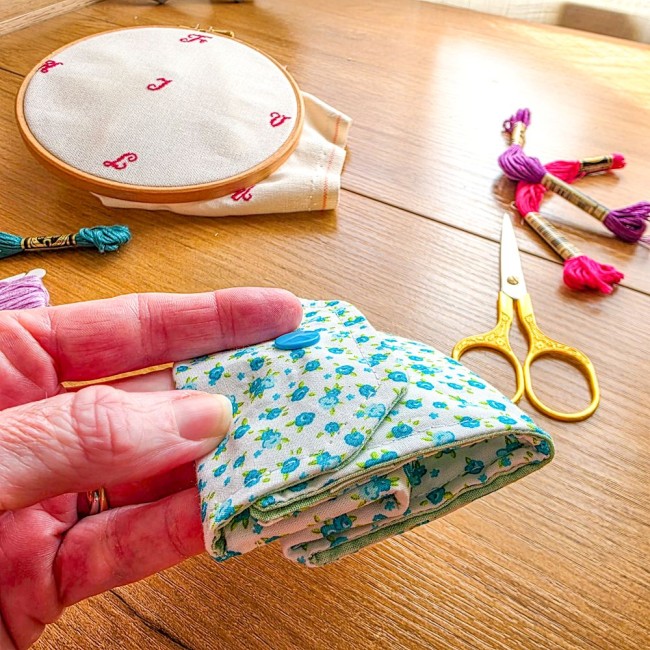 poubelle-a-fils-mini-pochon-point-de-croix-myosotis-plie-plat-angelique-la-fee-en-fil Gros plan d'une mini poubelle à fils pliable imprimée de fleurs de myosotis bleues tenue pliée à plat dans la main pour montrer sa finesse, posée au-dessus d'une table en bois avec du matériel de broderie, création artisanale Angélique La Fée En Fil.