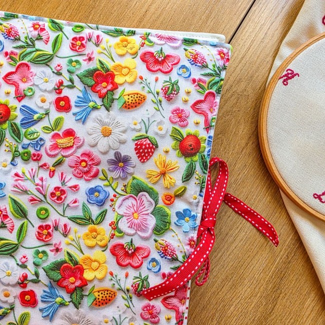 trousse-brodeuse-fleurs-fraises-alice-angelique-la-fee-en-fil Vue rapprochée de la trousse de brodeuse Alice, motif brodé fleurs et fraises, avec son nœud en ruban rouge et un tambour à broder sur une table en bois.