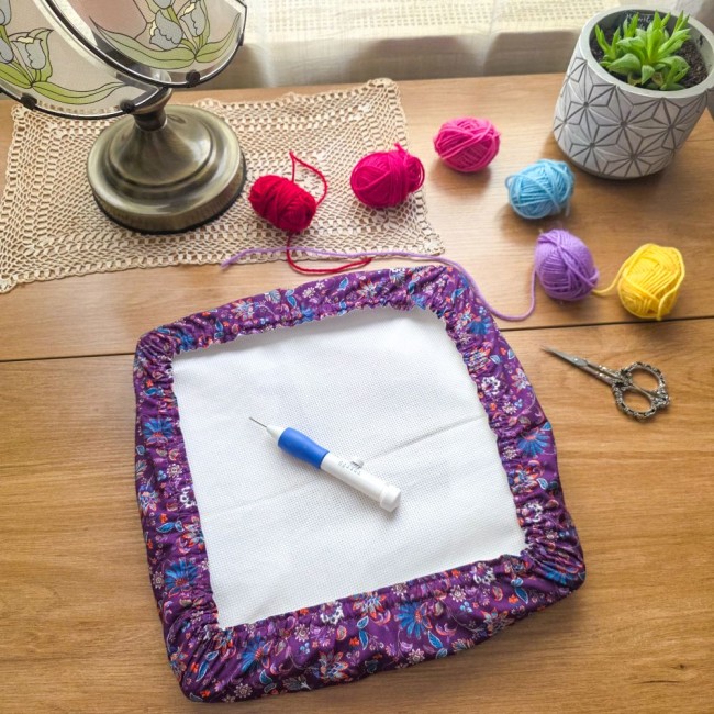 Vue de dessus d'un grime guard housse protège-toile modèle Cachemire en tissu violet aux motifs floraux et cachemire multicolores, installé sur un cadre carré avec un stylo punch needle, des pelotes de laine colorées et ciseaux, création artisanale Angéli
