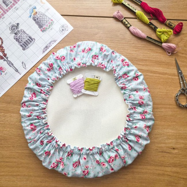 Vue de dessus d'un grime guard rond housse protège-toile modèle Bianca installé sur un tambour à broder rond, en tissu gris clair aux motifs bucoliques de roses rouges, avec carte de fils, ciseaux ciselés, échevettes et grille de point de croix sur une ta