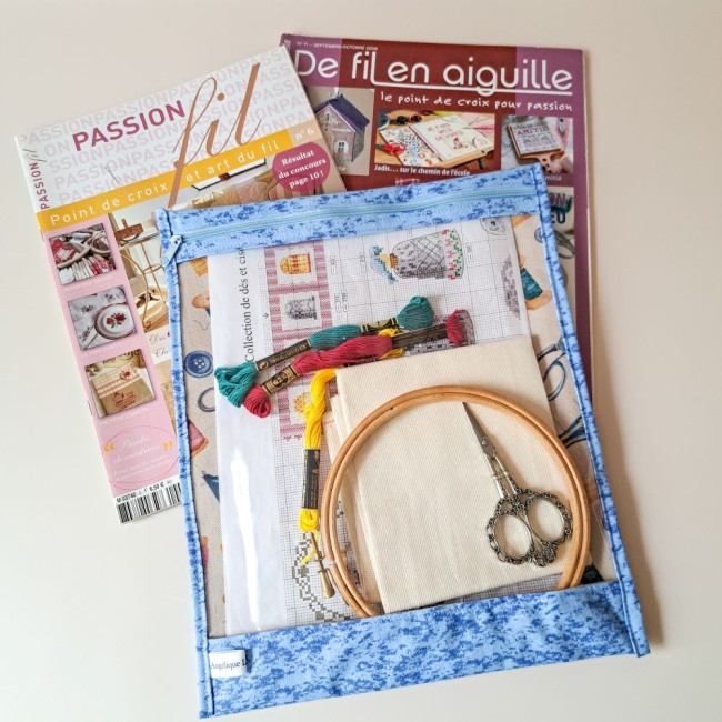 Posé sur la table une pochette à projet de broderie à l'intérieur des échevettes de fils à broder, un tambour à broder, une grille de point compté, une toile à broder. A côté 2 magazines de point de croix. Une création Angélique La Fée En Fil