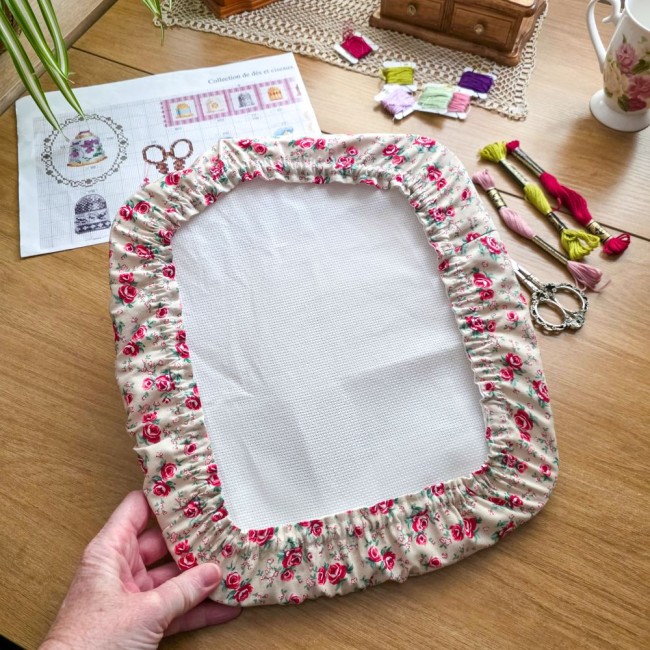 Gros plan des mains tenant un grime guard housse protège-toile modèle Vanille installé sur un grand cadre à broder rectangulaire, en tissu écru vanille aux motifs bucoliques de roses roses et rouges, avec ciseaux vintage, échevettes et grille de point de 