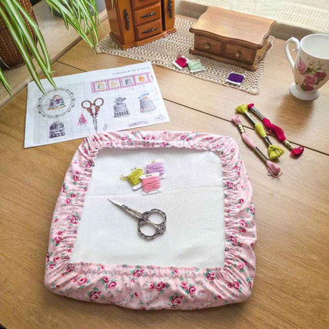 grime-guard-housse-protege-toile-broderie-romane-angelique-la-fee-en-fil Vue d'ensemble d'un grime guard housse protège-toile modèle Romane installé sur un cadre à broder carré, en tissu rose aux motifs bucoliques de fleurs et roses, posé sur une table en bois avec diagramme de broderie, ciseaux rétro et échevettes de fils, cr