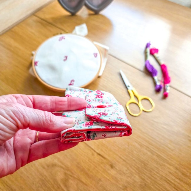 poubelle-a-fils-mini-pochon-broderie-vanille-pliee-main-angelique-la-fee-en-fil Gros plan de la mini poubelle à fils pliable fermée modèle Vanille tenue en main, montrant sa finesse et sa taille compacte en tissu écru effet lin aux motifs bucoliques de roses rouges, avec tambour à broder, ciseaux dorés et échevettes en arrière-plan,