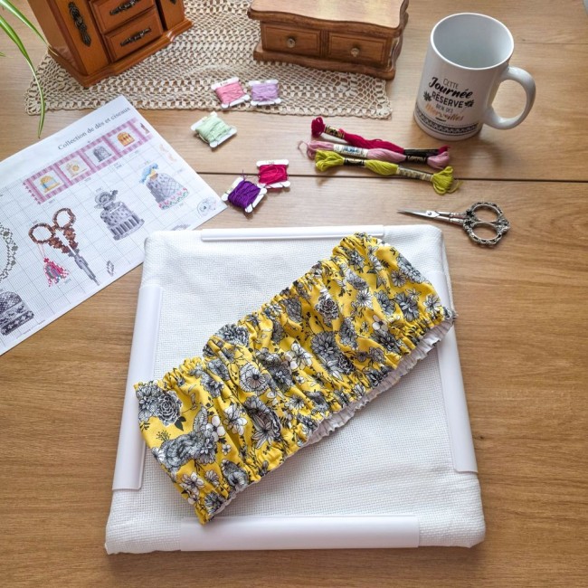 Grime guard modèle Julie en tissu jaune floral plissé, posé au centre d'un métier Q-Snap avec sa toile à broder blanche sur une table en bois par Angélique La Fée en Fil.
