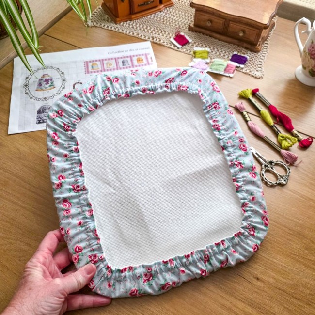 Gros plan des mains tenant un grime guard housse protège-toile modèle Bianca installé sur un grand cadre à broder rectangulaire, en tissu gris clair aux motifs bucoliques de roses rouges, avec ciseaux vintage, échevettes et grille de point de croix en arr