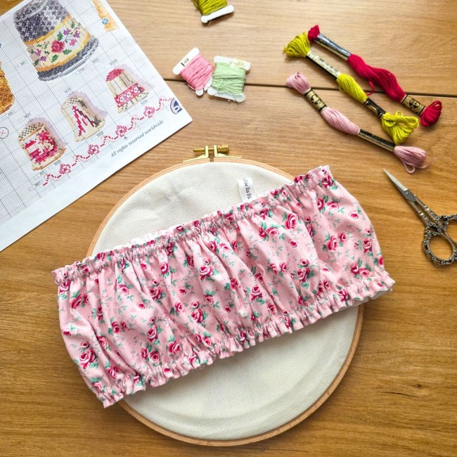 grime-guard-tambour-bois-broderie-romane-angelique-la-fee-en-fil Vue d'ensemble d'un grime guard housse protège-toile modèle Romane posé en travers sur un tambour à broder rond en bois, en tissu rose aux motifs bucoliques de fleurs et roses avec étiquette de marque, entouré d'une grille de broderie, échevettes et cisea