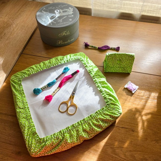 Vue de dessus d'un ensemble coordonné de broderie modèle Verveine comprenant un grime guard installé sur cadre Q-Snap carré et sa poubelle à fils de table assortie, en tissu vert anis aux motifs de feuillage blanc, avec ciseaux dorés, échevettes et boîte 