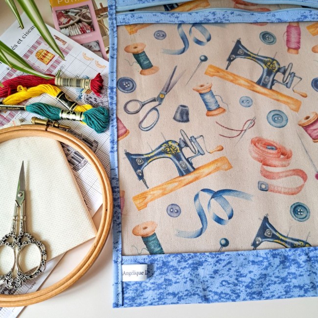  Posé sur une table, un sac à projet de broderie bleu motif couture. A côté des accessoires de broderie, tambour à broder, ciseaux à broder, toile à broder, des echevettes et une grille de point compté. Création Angélique La Fée En Fil
