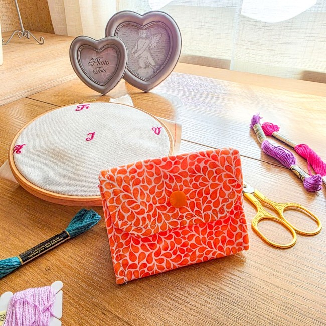 Mini poubelle à fils de table pliante modèle Mandarine en tissu coton orange à motifs gouttelettes blanches présentée fermée, avec un tambour à broder, des ciseaux dorés et des échevettes sur une table en bois, création artisanale Angélique La Fée En Fil.