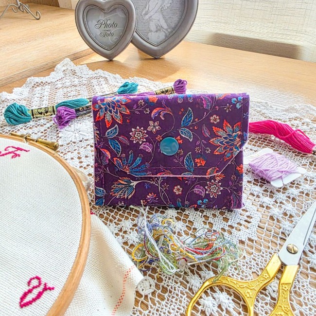 Mini poubelle à fils pliable en tissu violet imprimé de motifs cachemire et fleurs multicolores avec bouton-pression bleu turquoise, posée sur un napperon en dentelle avec accessoires de broderie, création artisanale Angélique La Fée En Fil.