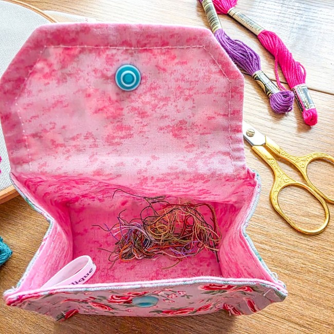 Vue plongeante de l'intérieur d'une mini poubelle à fils ouverte modèle Bianca, montrant les chutes de fils de broderie au fond du pochon rose pastel avec étiquette de marque, entourée de ciseaux dorés et d'échevettes, création artisanale Angélique La Fée