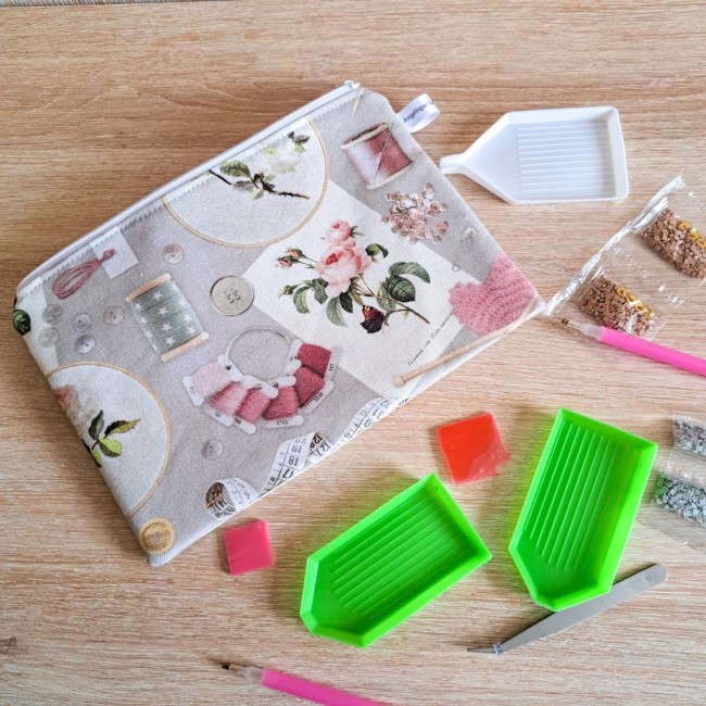 trousse-zippee-motif--broderie-diamant broderie-angelique-la-fee-en-fil pochette zippée en coton motif accessoire de point de croix posé sur la table, près d'elle des barquettes de broderie diamant, de la colle et des stylets