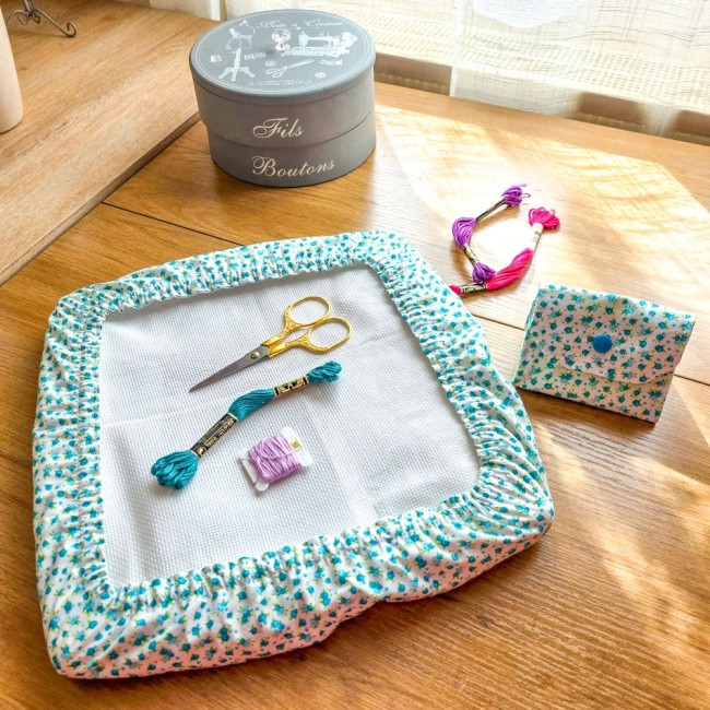 poubelle-a-fils-couvre-tambour-assorti-myosotis-broderie-angelique-la-fee-en-fil Ensemble d'accessoires de broderie assortis modèle Myosotis comprenant une mini poubelle à fils pliable et une housse protège-toile pour tambour carré en tissu floral bleu, accompagnés de ciseaux dorés, d'échevettes et d'une boîte à couture grise, créatio