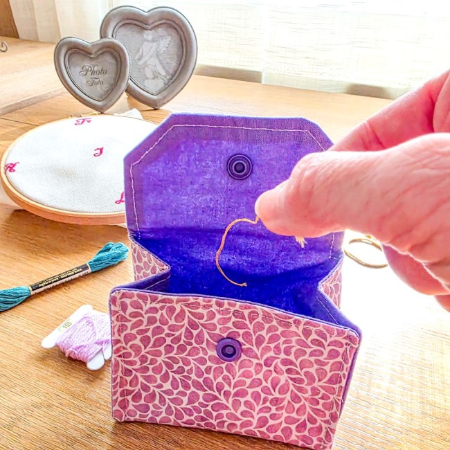Gros plan sur l'utilisation d'une mini poubelle à fils de table dépliante ouverte, modèle Lavande en tissu coton violet à motifs feuillage et doublure intérieure violet uni, avec une main y déposant un brin de fil, sur fond d'accessoires de broderie, créa