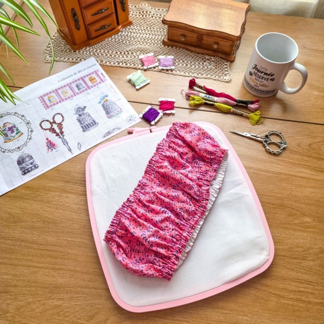 grime-guard-sous-bois-rose-sur-cadre-nurge-rose-angelique-la-fee-en-fil Un couvre-cadre (grime guard) élastiqué modèle Sous-bois Rosé en coton vieux rose à motifs de branches violettes, posé en diagonale sur une toile Aïda blanche tendue dans un cadre carré rose de marque Nurge. La mise en scène sur une table en bois comprend