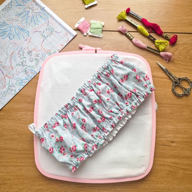 Gros plan des mains tenant un grime guard housse protège-toile modèle Bianca installé sur un grand cadre à broder rectangulaire, en tissu gris clair aux motifs bucoliques de roses rouges, avec ciseaux vintage, échevettes et grille de point de croix en arr