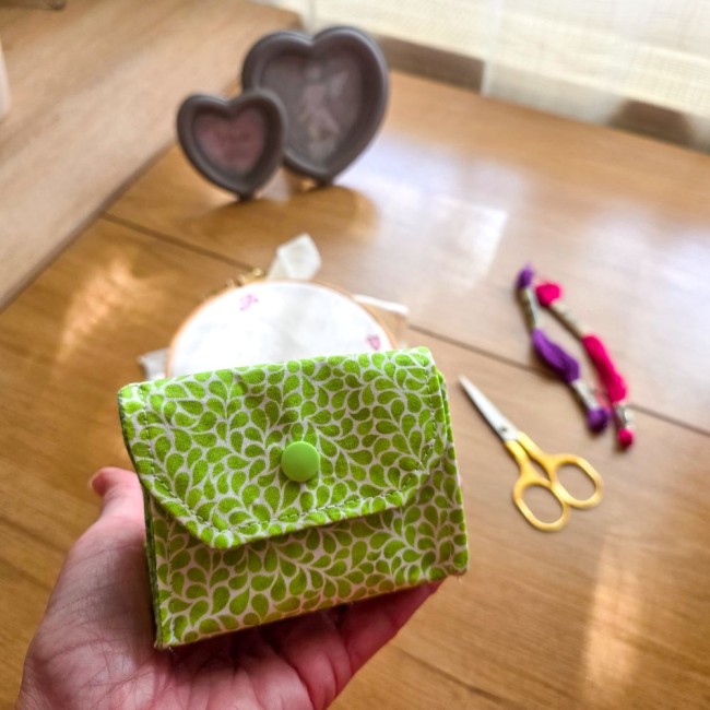 Gros plan de la main tenant une mini poubelle à fils modèle Verveine avec pression verte assortie, en tissu vert anis aux motifs de feuillage blanc, avec ciseaux dorés et échevettes de fils en arrière-plan, création artisanale Angélique La Fée En Fil.