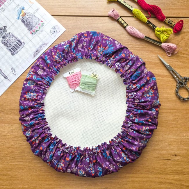 Vue de dessus d'un grime guard rond modèle Cachemire en tissu violet aux motifs floraux et cachemire multicolores, installé sur un tambour à broder rond avec ciseaux brodeuse, échevettes de fils et grille de point de croix, création artisanale Angélique L