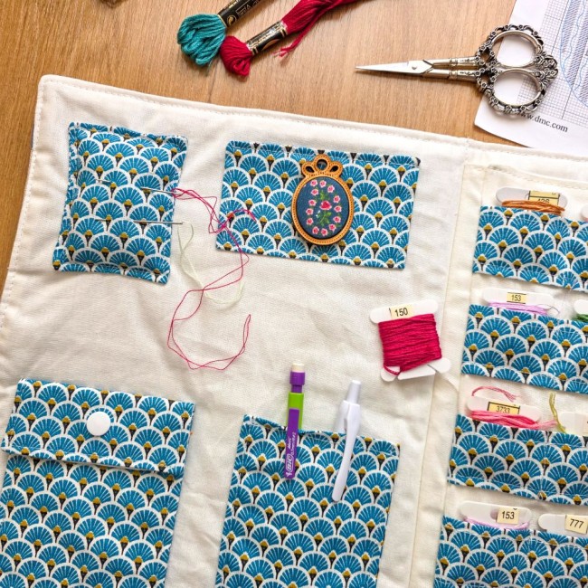 trousse-brodeuse-pique-aiguille-rangement-accessoires-mains-et-merveilles-angelique-la-fee-en-fil Vue de l'intérieur d'une trousse de brodeuse Mains et Merveilles ouverte. Elle est fabriquée en tissu crème avec des poches et un pique-aiguille en tissu bleu à motif éventail Art Déco. La trousse est remplie d'accessoires : aiguilles piquée, un dé en boi