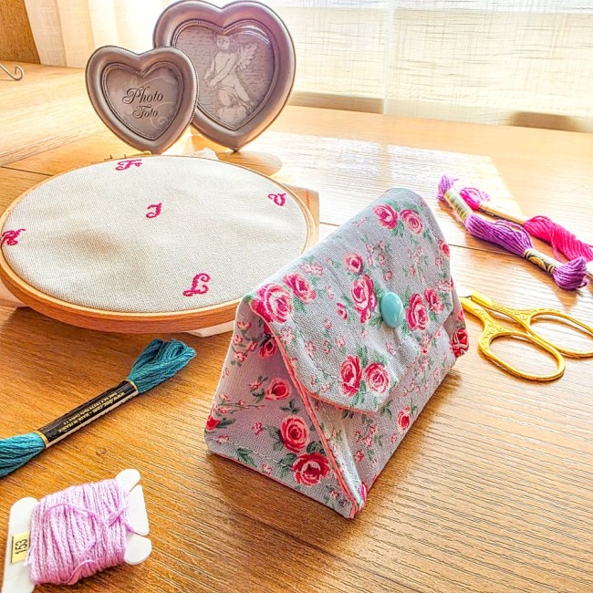 Vue de trois-quarts d'une mini poubelle à fils pliable fermée pour broderie et point de croix, modèle Bianca en tissu gris clair aux motifs bucoliques de roses rouges avec rabat et bouton-pression bleu clair, posée sur une table en bois à côté d'un tambou