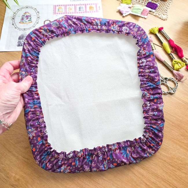 Gros plan sur la prise en main d'un grime guard modèle Cachemire en tissu violet aux motifs floraux et cachemire multicolores, ajusté sur un cadre Q-Snap carré avec échevettes de fils et grille de broderie en arrière-plan, création artisanale Angélique La