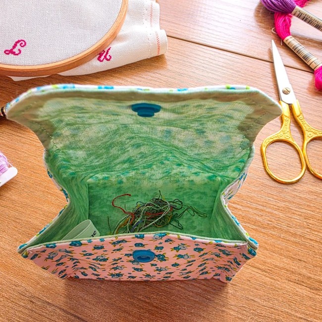 poubelle-a-fils-mini-pochon-point-de-croix-myosotis-interieur-brins-de-fils-angelique-la-fee-en-fil Vue plongeante de l'intérieur d'une mini poubelle à fils pliable ouverte en tissu écru imprimé myosotis bleus avec doublure verte contenant des chutes de fils, posée sur une table en bois à côté de ciseaux dorés et d'un tambour, création artisanale Angéli