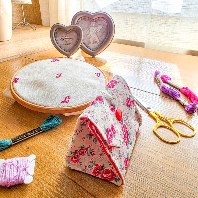poubelle-a-fils-point-de-croix-vanille-trois-quarts-angelique-la-fee-en-fil Vue de trois-quarts d'une mini poubelle à fils pliable fermée pour broderie et point de croix, modèle Vanille en tissu écru au rendu naturel effet lin imprimé de motifs bucoliques de roses rouges avec rabat et bouton-pression rouge vif, posée sur une tabl