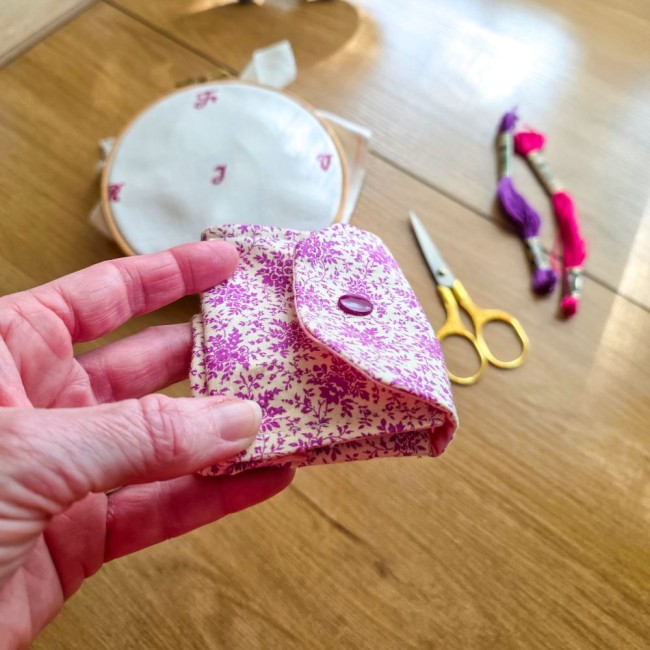 Vue de profil d'une mini poubelle à fils de table pliante modèle Samba en tissu coton à motifs floraux rose et violet tenue en main, avec un tambour à broder, des ciseaux dorés et des échevettes en arrière-plan, création artisanale Angélique La Fée En Fil