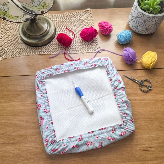 Vue d'ensemble d'un grime guard housse protège-toile modèle Bianca utilisé pour le punch needle sur cadre carré, en tissu gris clair aux motifs bucoliques de roses rouges, posé sur une table en bois avec stylo punch needle, pelotes de laine colorées et ci