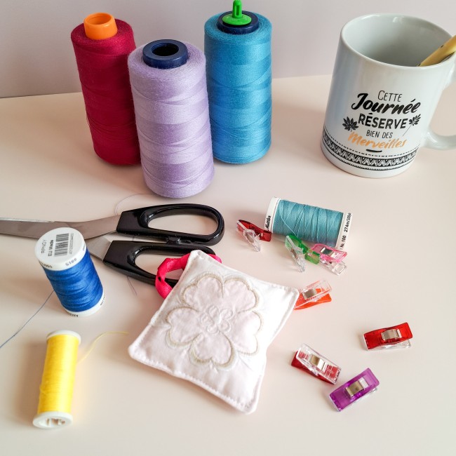 bijou-ciseaux-brodeuse-coussinet-fleur-blanche-fils-bobines-pinces-atelier-couture-angelique-la-fee-en-fil.jpg Bijou de ciseaux pour brodeuse en forme de coussinet blanc brodé d’une fleur, entouré de bobines de fil colorées, de pinces de couture, d’une paire de ciseaux et d’une tasse, création artisanale Angélique La Fée En Fil.