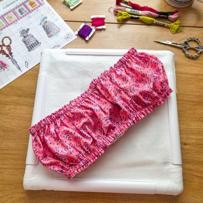grime-guard-sous-bois-rose-sur-cadre-qsnap-blanc-angelique-la-fee-en-fil Un couvre-cadre (grime guard) élastiqué modèle Sous-bois Rosé en coton vieux rose à motifs de branches violettes, posé en diagonale sur une toile Aïda blanche fixée sur un cadre rectangulaire blanc de type Q-Snap. La mise en scène sur une table en bois co
