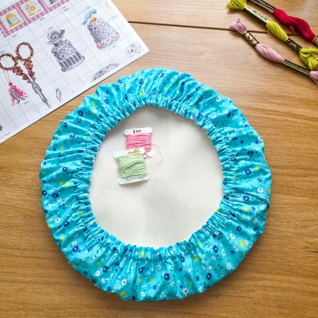 grime-guard-tambour-rond-bleu-turquoise-fleuri-angelique-la-fee-en-fil Un couvre-tambour (grime guard) rond en coton bleu turquoise imprimé de petites fleurs blanches, bleues et jaunes, installé sur un cercle à broder avec sa toile blanche. Deux cartonnettes de fils (rose et vert d'eau) sont posées au centre. La mise en scèn