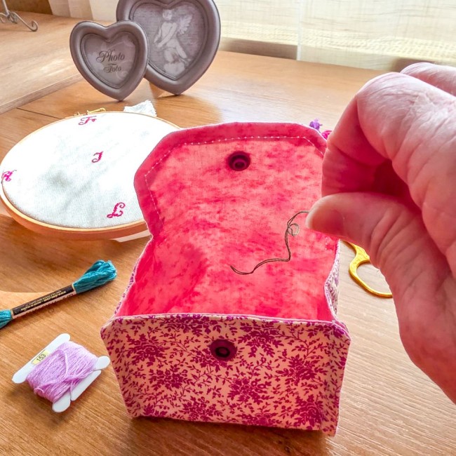 Vue rapprochée d'une mini poubelle à fils de table ouverte modèle Samba en tissu coton à motifs floraux rose et violet, montrant l'intérieur uni contrasté pendant qu'une main y dépose des fils coupés, avec un tambour à broder en arrière-plan, création art
