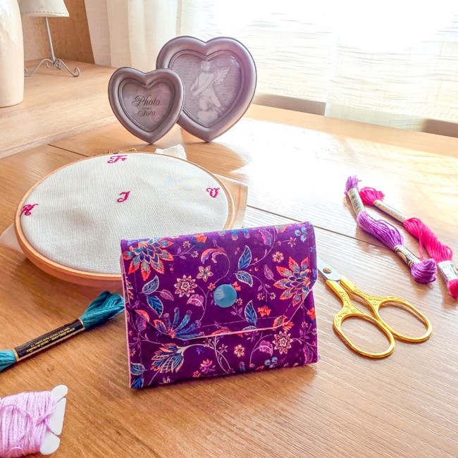 Vue de face d'une mini poubelle à fils de table fermée modèle Cachemire en tissu violet aux motifs floraux et cachemire multicolores, avec bouton-pression bleu, ciseaux dorés et tambour à broder en arrière-plan, création artisanale Angélique La Fée En Fil