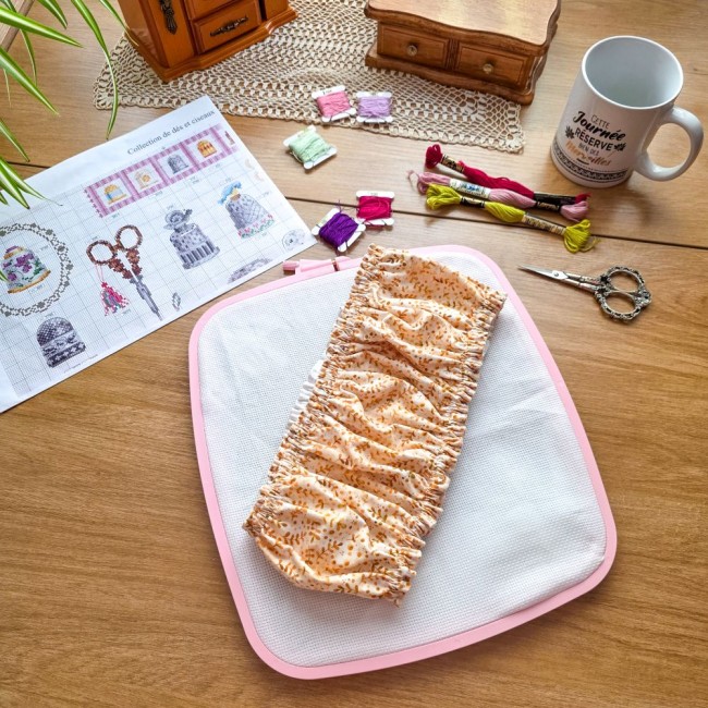 grime-guard-broderie-sous-bois-dore-tissu-beige-replie-tambour-nurge-rose-angelique-la-fee-en-fil Grime guard modèle Sous-Bois Doré en tissu beige plissé à motifs de feuillage ocre, posé au centre d'un tambour à broder rectangulaire Nurge rose sur un bureau d'atelier par Angélique La Fée en Fil.