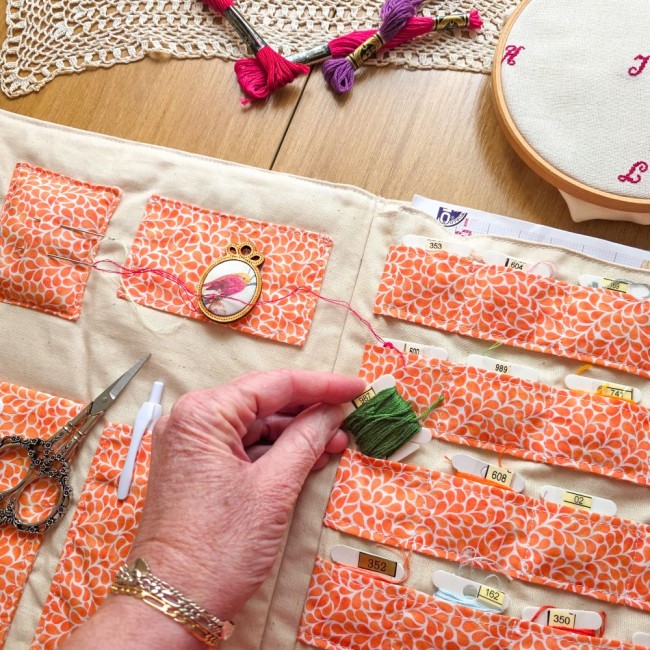 Main attrapant une cartonnette de fil vert dans les rangements de la trousse de brodeuse Savannah en tissu orange à motif gouttes, création artisanale Angélique La Fée En Fil.