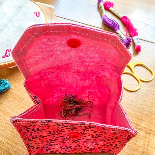 Vue plongeante de l'intérieur d'une mini poubelle à fils ouverte modèle Sandy Rose, montrant les chutes de fils de broderie au fond du pochon rose vif, entourée de ciseaux dorés et d'échevettes de fils, création artisanale Angélique La Fée En Fil.