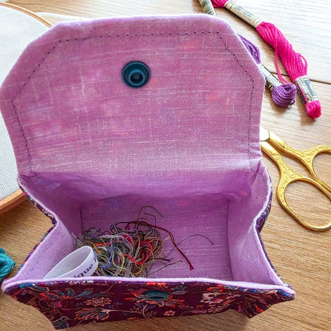 Gros plan sur l'intérieur d'une mini poubelle à fils nomade et pliante modèle Cachemire ouverte avec sa doublure en tissu rose, contenant des chutes de fils à broder, avec ciseaux dorés et échevettes en arrière-plan, création artisanale Angélique La Fée E