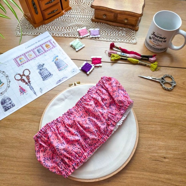 grime-guard-sous-bois-rose-sur-tambour-bois-angelique-la-fee-en-fil Un couvre-cadre (grime guard) élastiqué modèle Sous-bois Rosé en coton vieux rose à motifs de branches violettes, posé en diagonale sur une toile Aïda blanche tendue sur un tambour à broder rond en bois. La mise en scène sur une table en bois clair compre