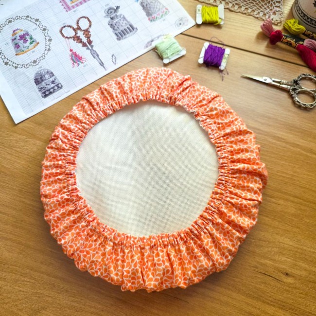 grime-guard-orange-tambour-a-broder-broderie-angelique-la-fee-en-fil Vue de dessus d'un grime guard housse protège-toile modèle Mandarine en tissu coton orange imprimé de motifs gouttelettes blanches, ajusté sur un tambour à broder rond, avec grille de broderie, ciseaux et échevettes sur une table en bois, création artisan
