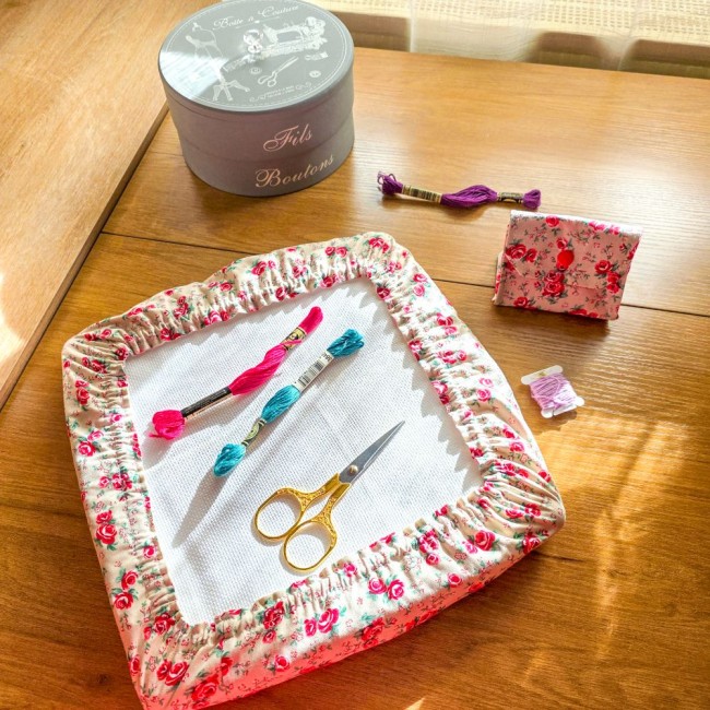 duo-broderie-grime-guard-poubelle-a-fils-vanille-angelique-la-fee-en-fil Ensemble d'accessoires de broderie assortis modèle Vanille comprenant un grime guard sur cadre à broder et une mini poubelle à fils pliée en tissu écru effet lin aux motifs bucoliques de roses rouges, disposés sur une table en bois avec ciseaux dorés et é