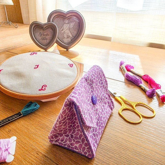 Vue de profil d'une mini poubelle à fils de table dépliante en position ouverte modèle Lavande en tissu coton violet à motifs feuillage, avec bouton-pression violet, tambour à broder, ciseaux dorés et échevettes de fils en arrière-plan, création artisanal